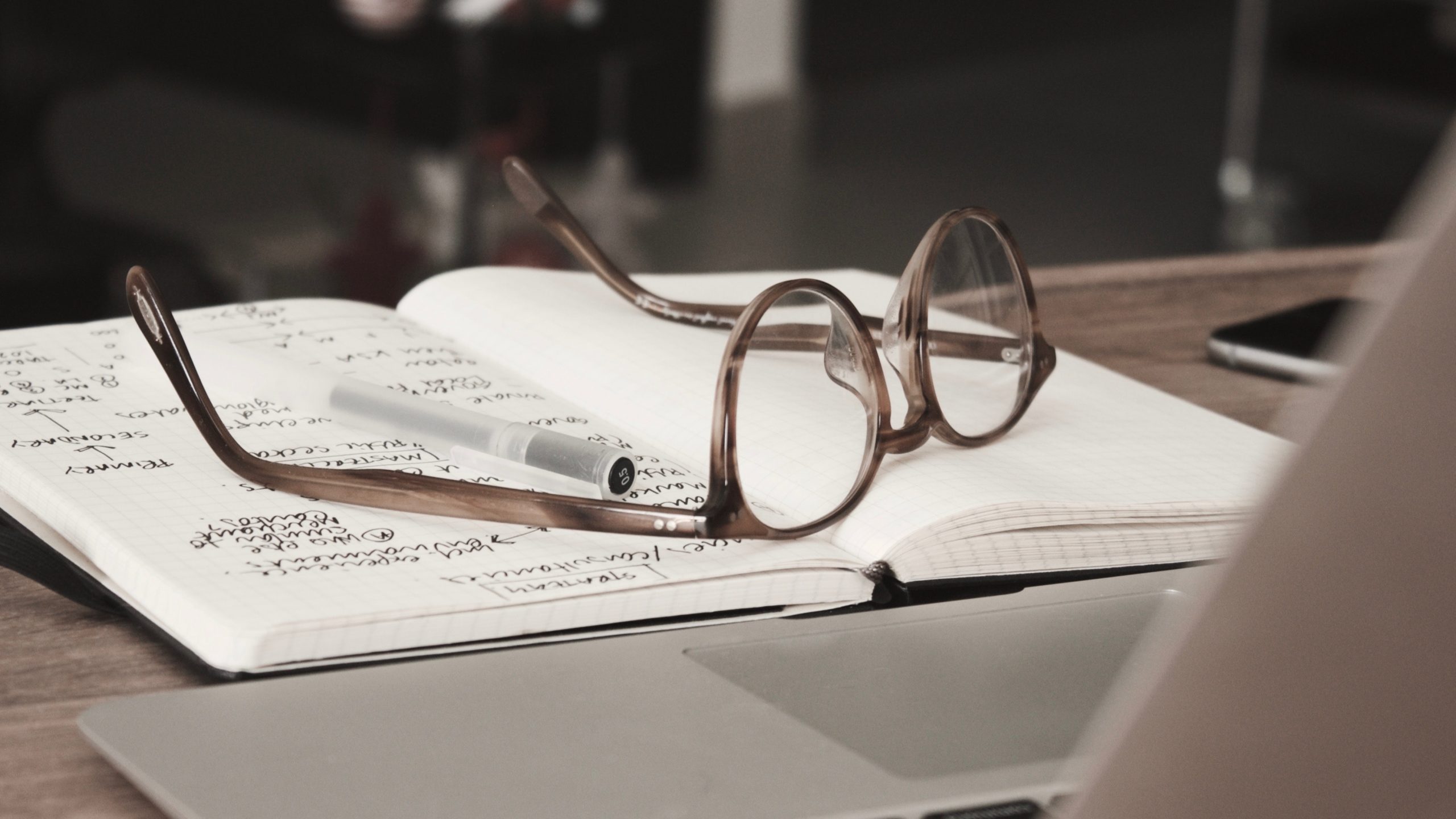 A pair of glasses and a pencil sit on an opened notebook in front of a laptop