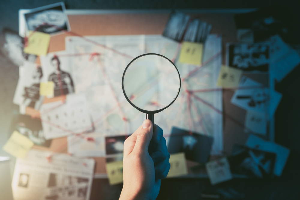 Detective hand holding a magnifying glass in front of a board with evidence crime scene photos and map high contrast image