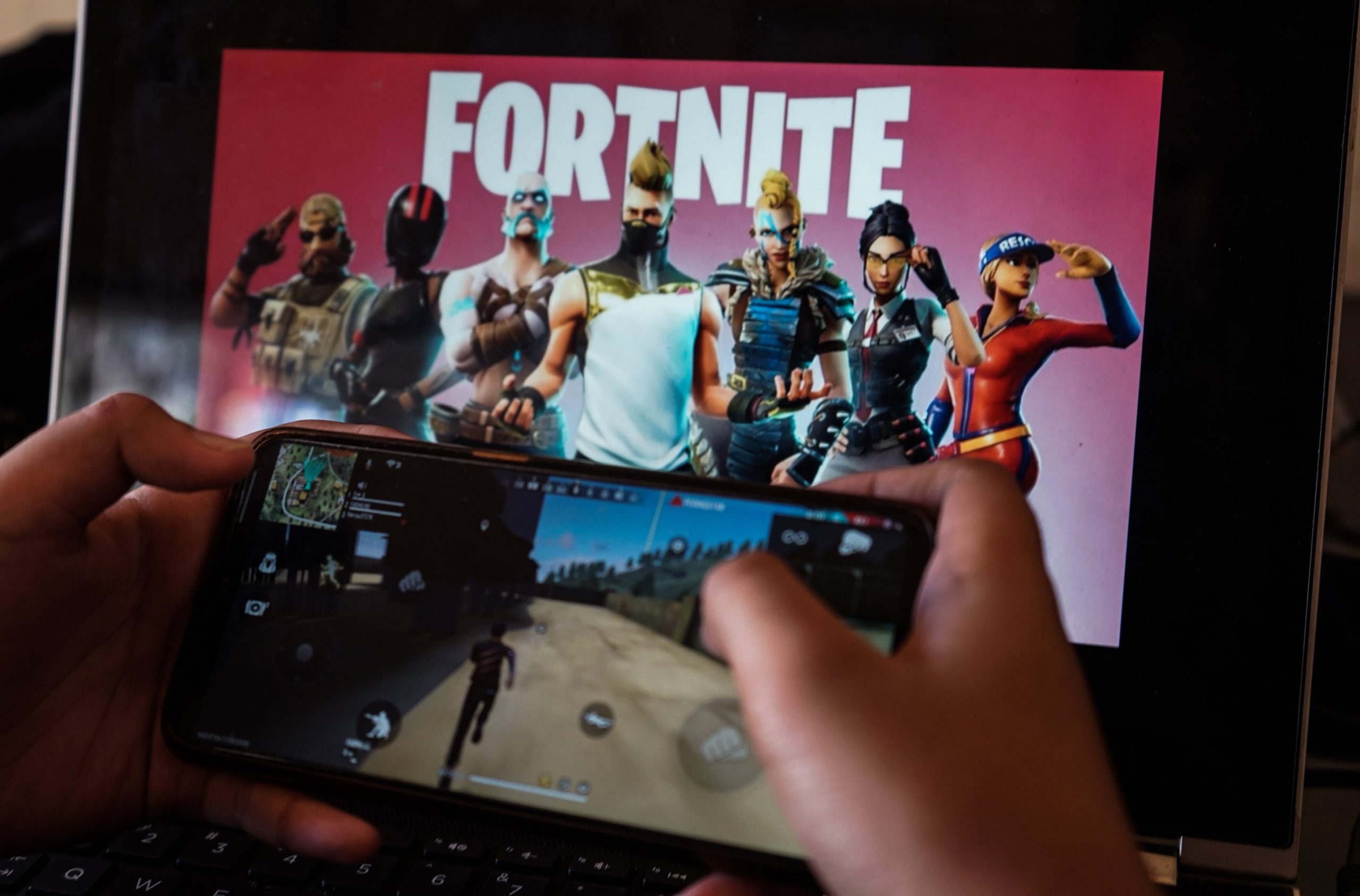 A child is playing a game on a cellphone with a picture of the Fortnite game on the computer screen in the background