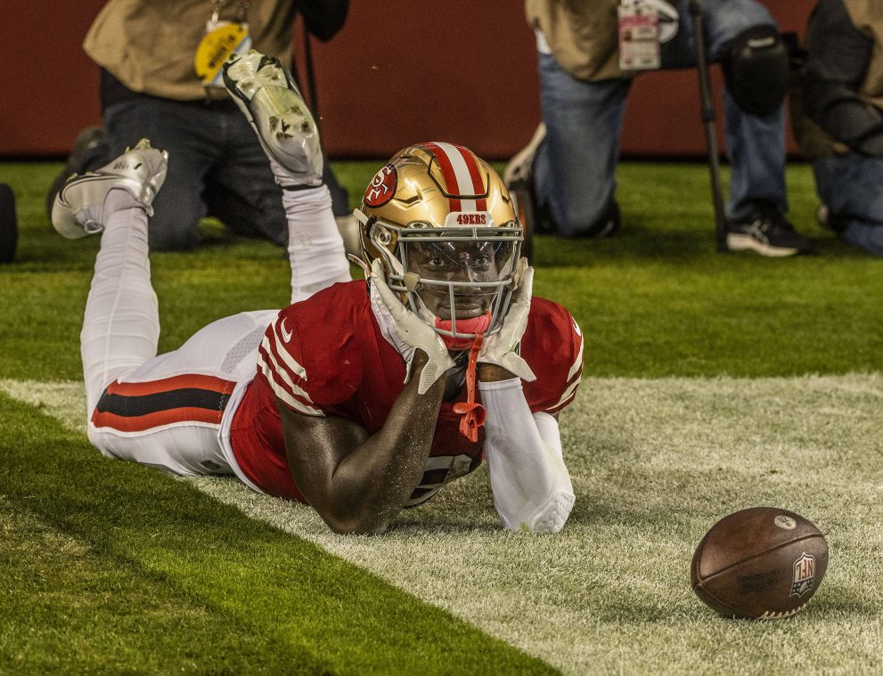 An american football player celebrates a touchdown sitting on the ground and looking at the ball